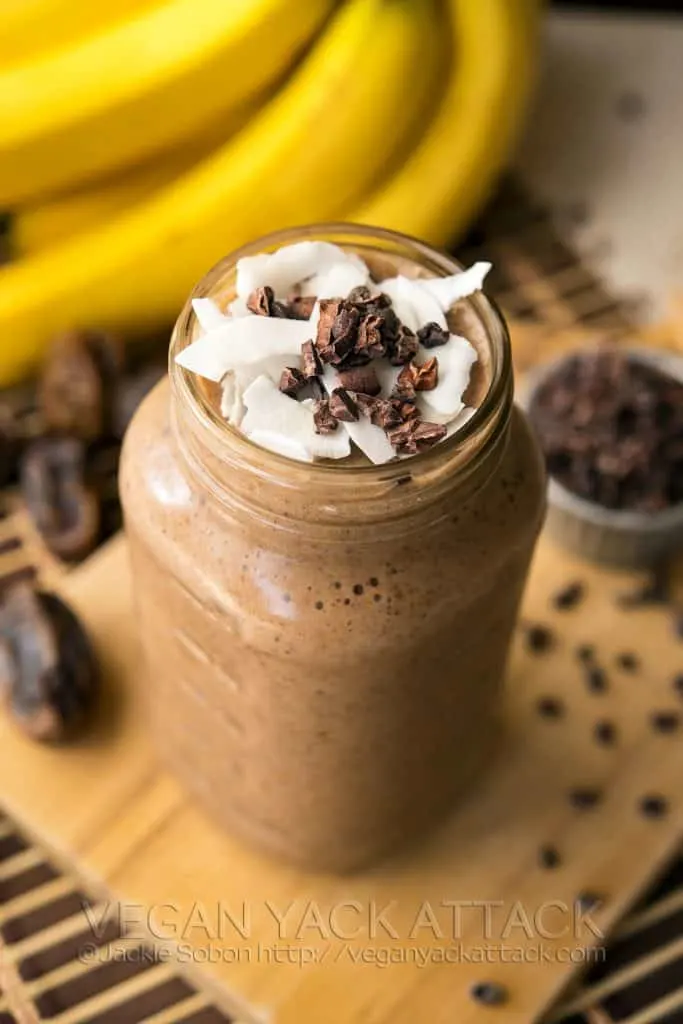 Image of chocolate banana smoothie in a jar on top of a cutting board, with bananas and dates in the background