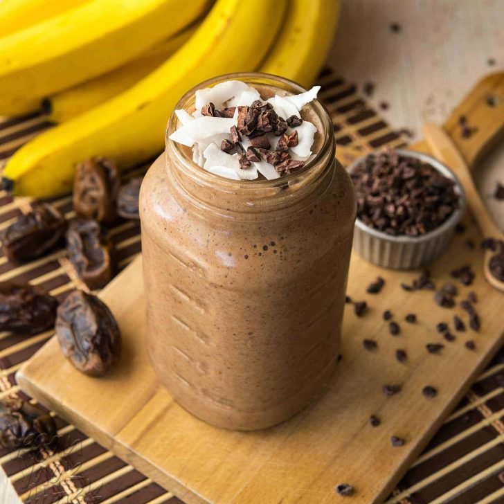 Large jar filled with banana cacao recovery smoothie, on a bread board surrounded by ingredients