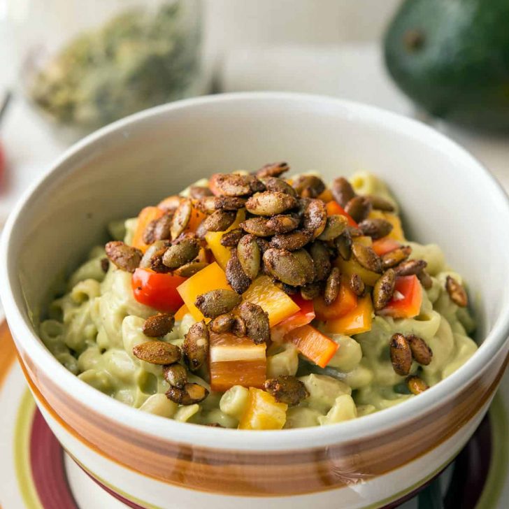 A striped bowl with Avocado Mac with Pepita Bacon inside, on a matching plate