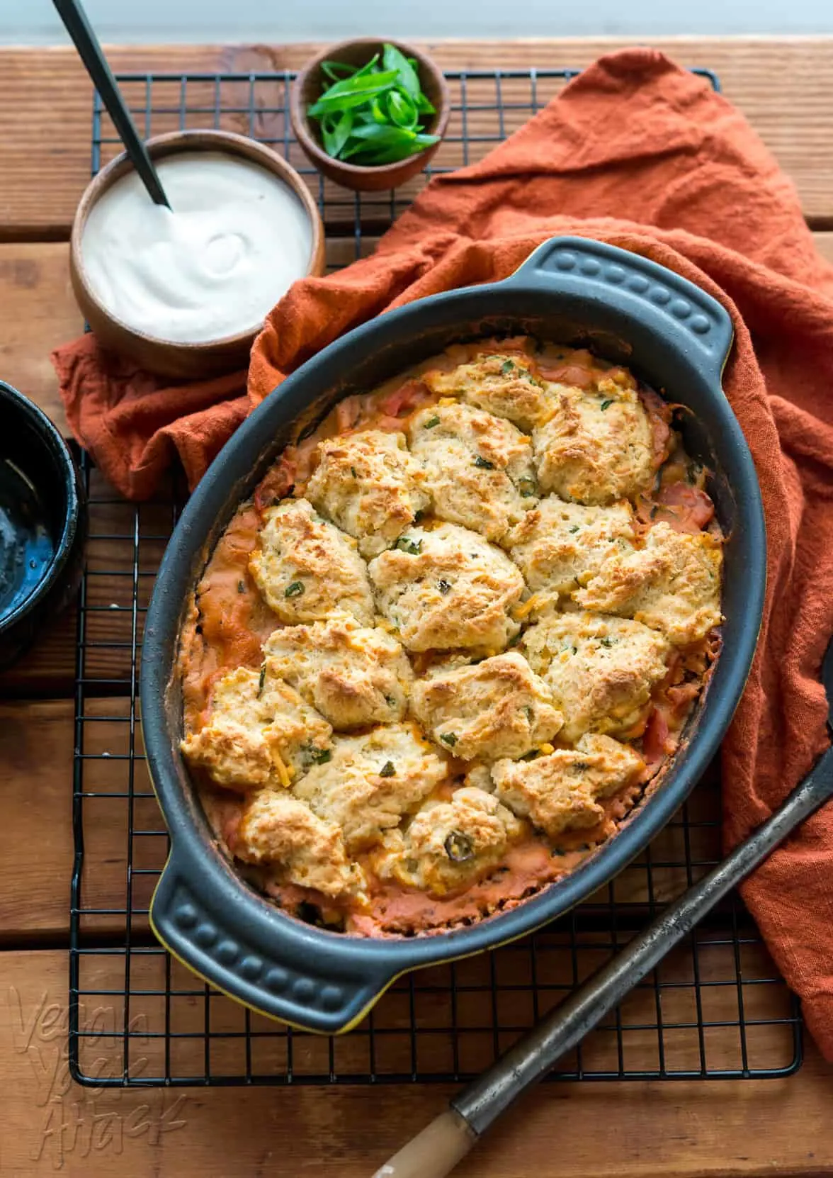 Tomato cobbler with biscuit topping in black oval dish with cooling rack and red linen