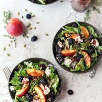 Three bowls of summery salad on a marble table top, next to seeds, a peach, and blackberries