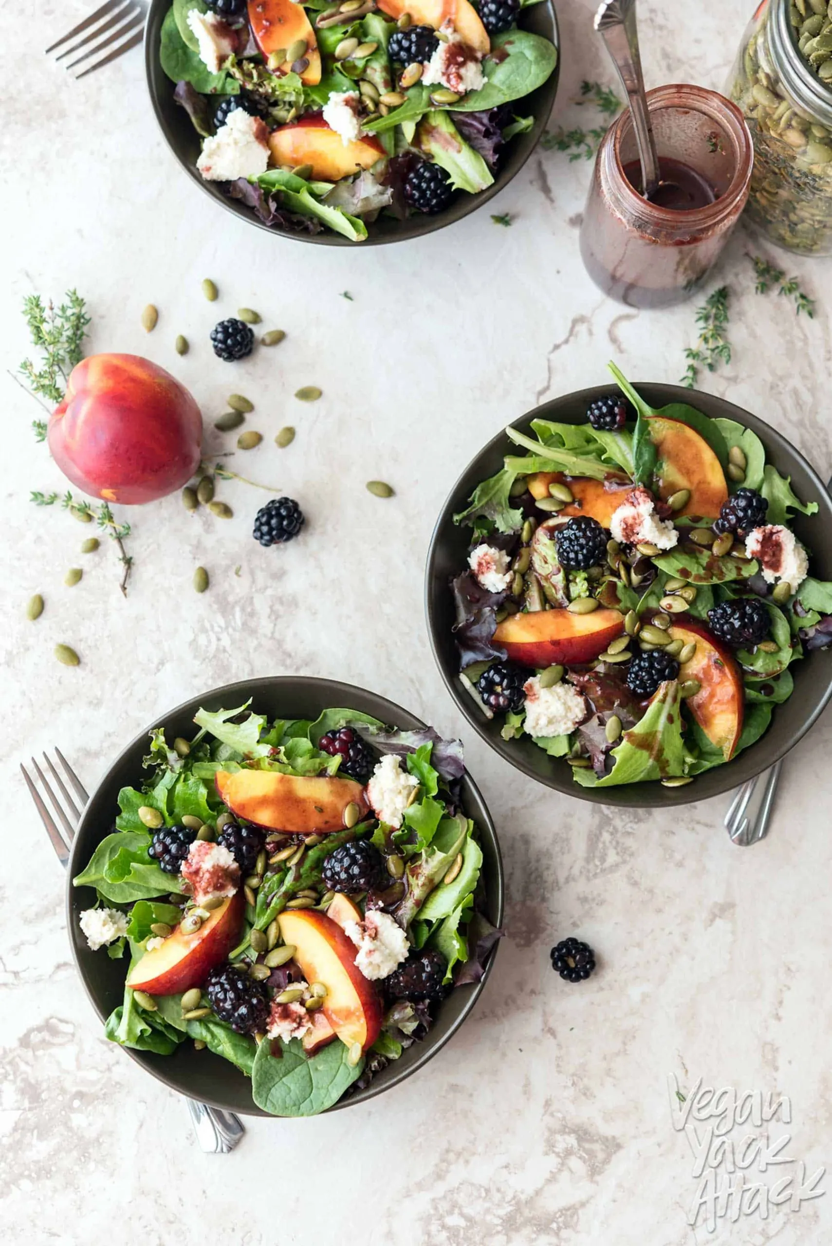 Three bowls of summery salad on a marble table top, next to seeds, a peach, and blackberries