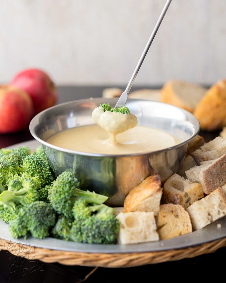 pale fondue dip in a metal bowl with chopped broccoli and bread around it