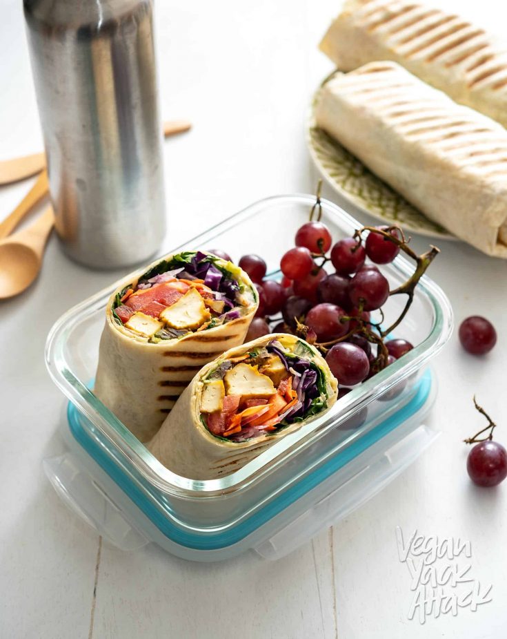 A chipotle tofu rainbow wrap cut in half in a lunch container with grapes