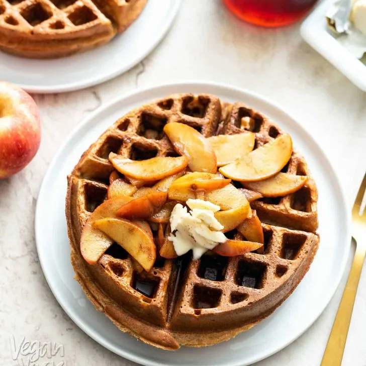 2 large, round, Belgian waffles on white plates, topped with apples and with syrup on the side