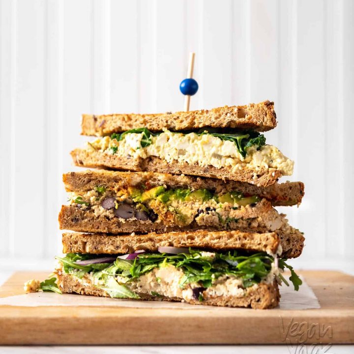 Image of three tofu 'egg' salad sandwiches stacked on one another on a cutting board