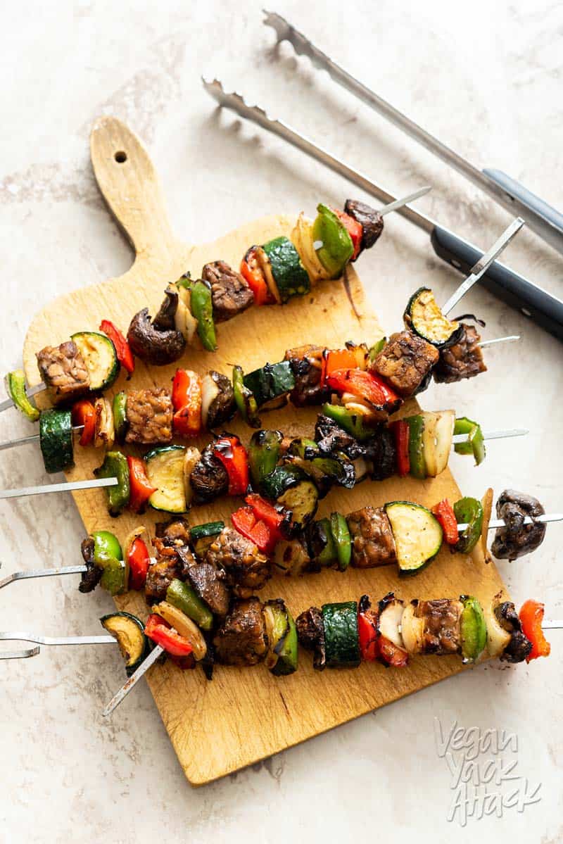 Tempeh veggie skewers stacked on a sandwich board with tongs next to them