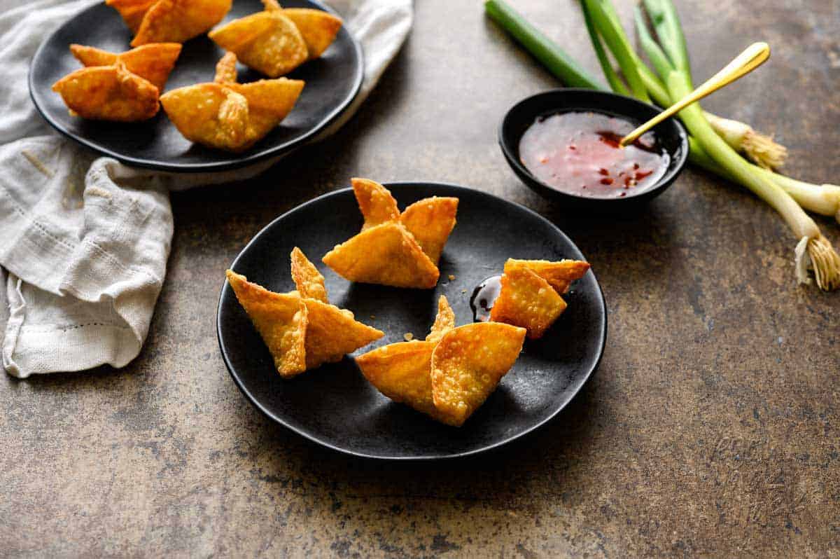 Vegan Crab Rangoon on black plates with a side of sweet chili sauce, whole green onions, and a white linen on a great background
