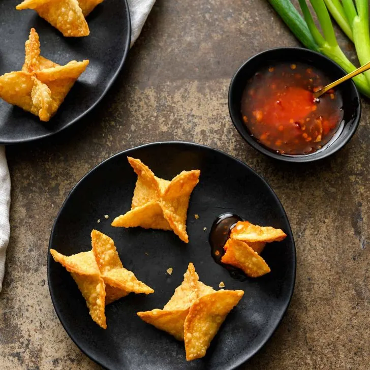 Vegan Crab Rangoon on black plates with a side of sweet chili sauce, whole green onions, and a white linen on a great background