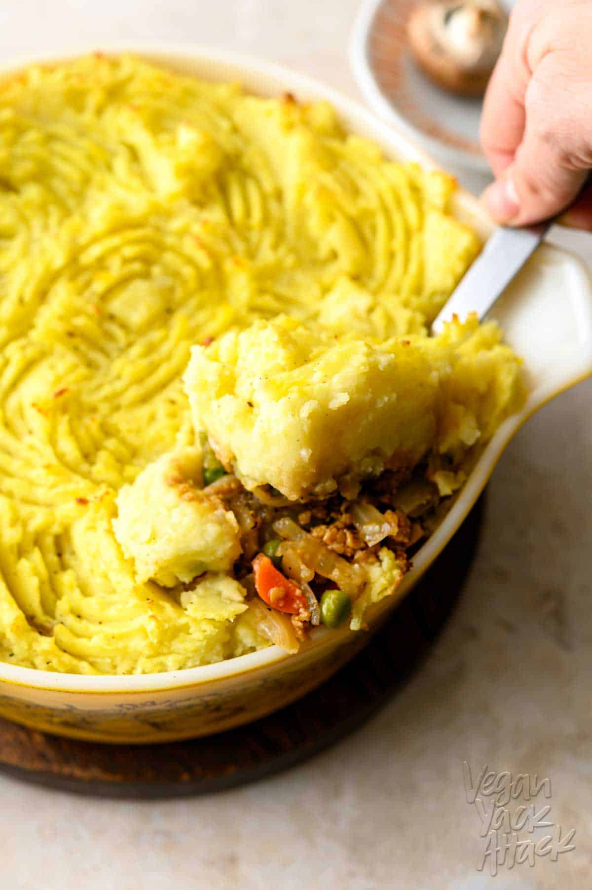 One casserole dish filled with a vegan shepherd's pie, being scooped out with a steel serving spoon, on a light linoleum background.