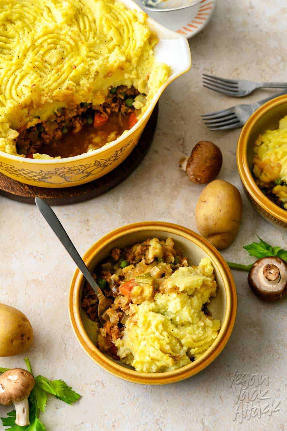 One casserole dish filled with a vegan shepherd's pie, spooned into two bowls, with potatoes and mushrooms scattered around them, on a light linoleum background.