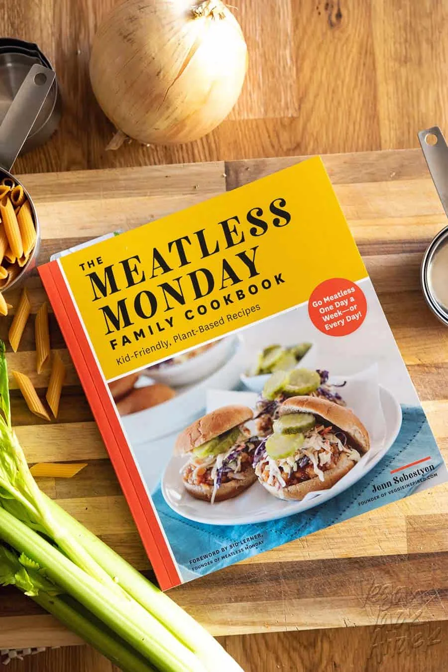 The Meatless Monday Family Cookbook on a cutting board with dried pasta, celery and onion.