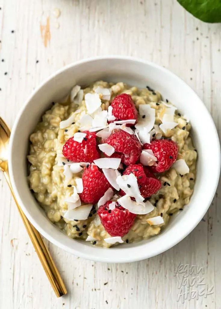 Image of white bowl with matcha oatmeal, topped with raspberries, on a white wood background