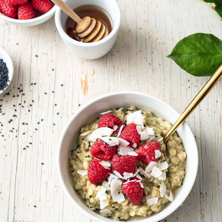Image of white bowl with matcha oatmeal, topped with raspberries, on a white wood background, with leaves to the side