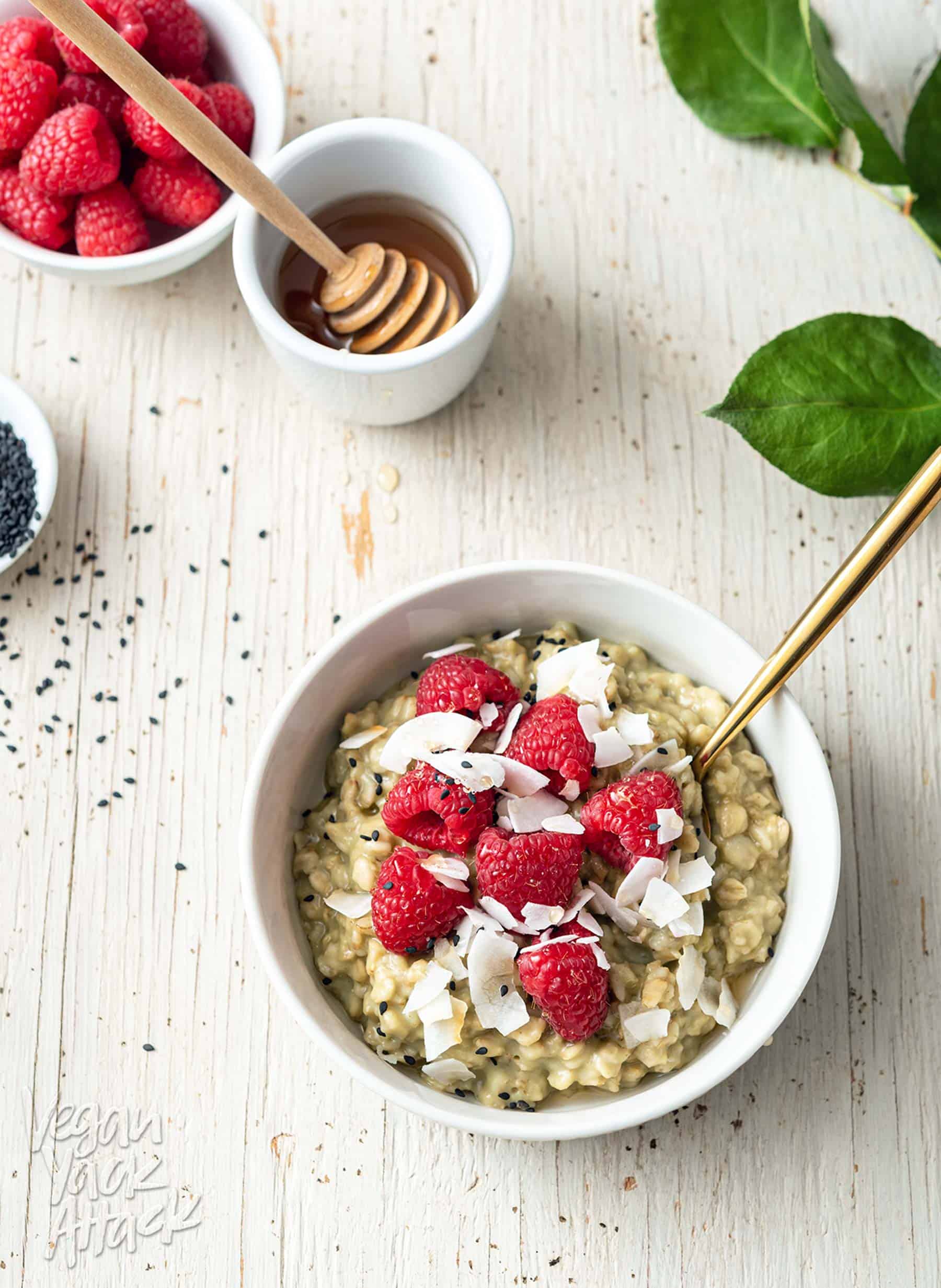 Image of white bowl with matcha oatmeal, topped with raspberries, on a white wood background, with leaves to the side