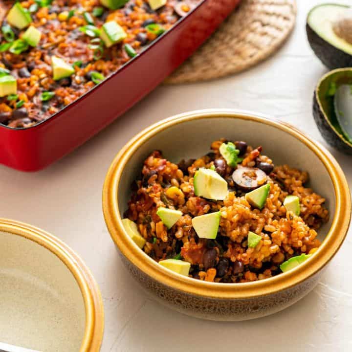 Image of chipotle brown rice bake in a red casserole dish on a beige background with bowl in foreground