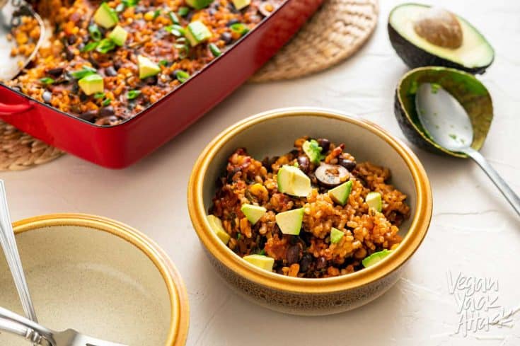 Image of chipotle brown rice bake in a red casserole dish on a beige background with bowl in foreground