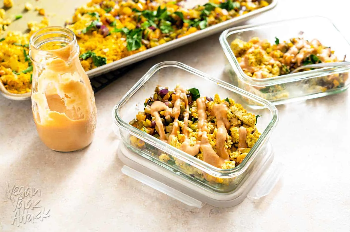 Two meal prep containers filled with Sheet Pan Tofu Scramble and hash, drizzled with sauce, next to a sheet pan