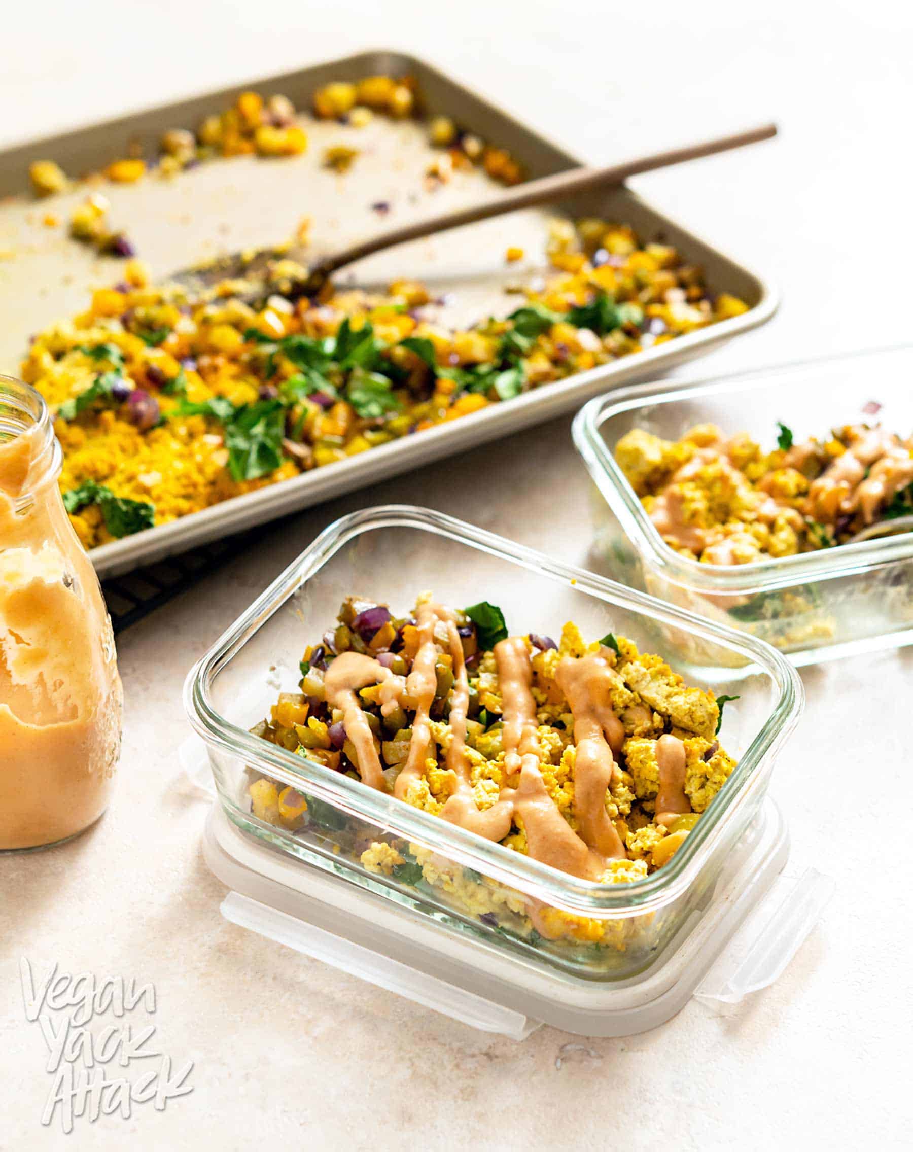 Two meal prep containers filled with Sheet Pan Tofu Scramble and hash, drizzled with sauce, next to a sheet pan