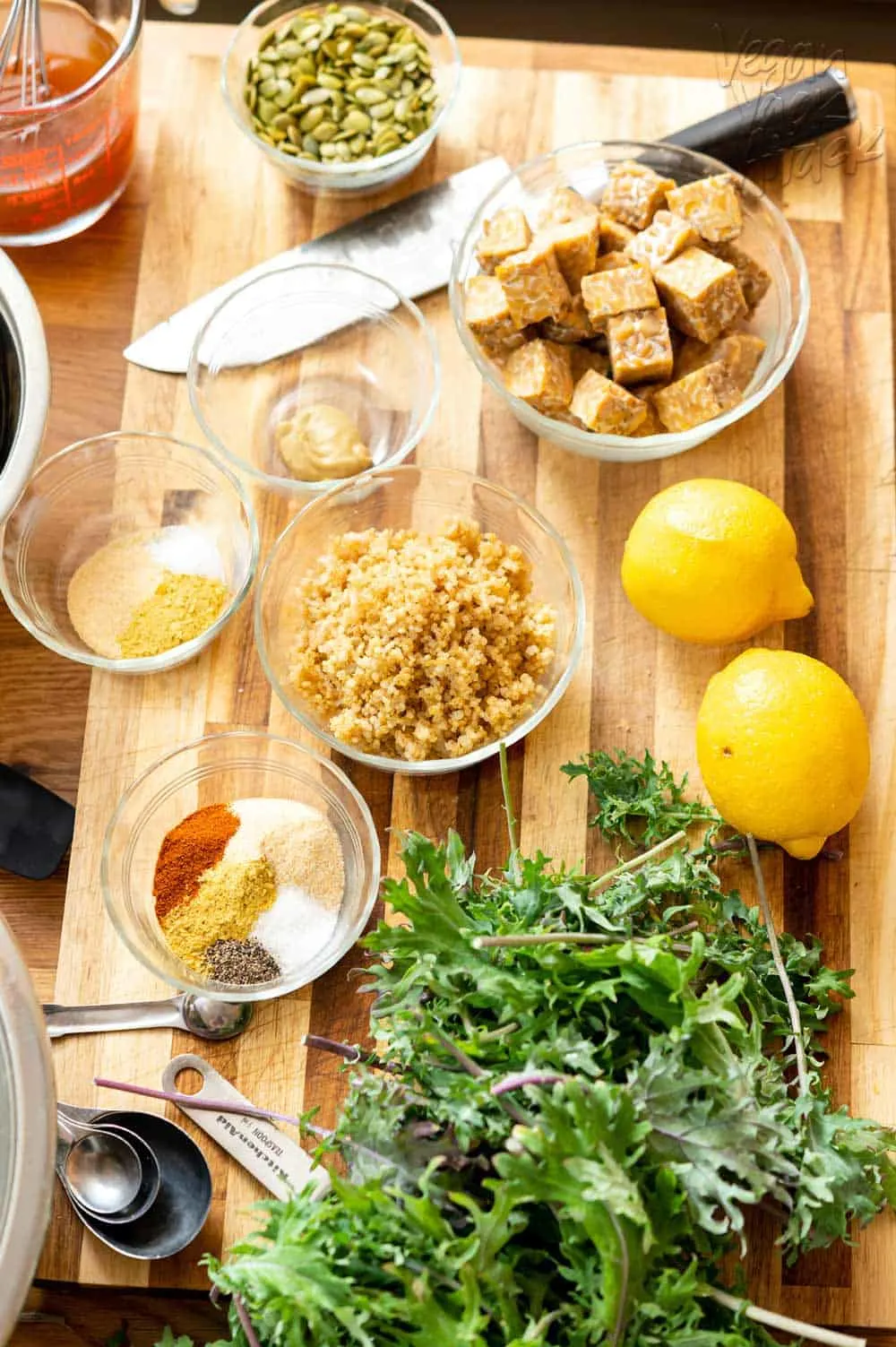 Ingredients such as tempeh, lemon, quinoa, spices, kale, and more laid out on a cutting board