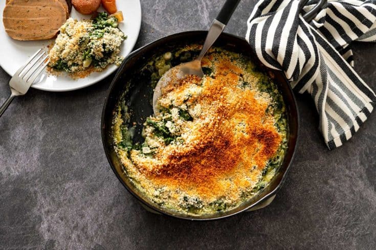 Serving spoon in a cast iron skillet filled with creamed kale next to dinner plates