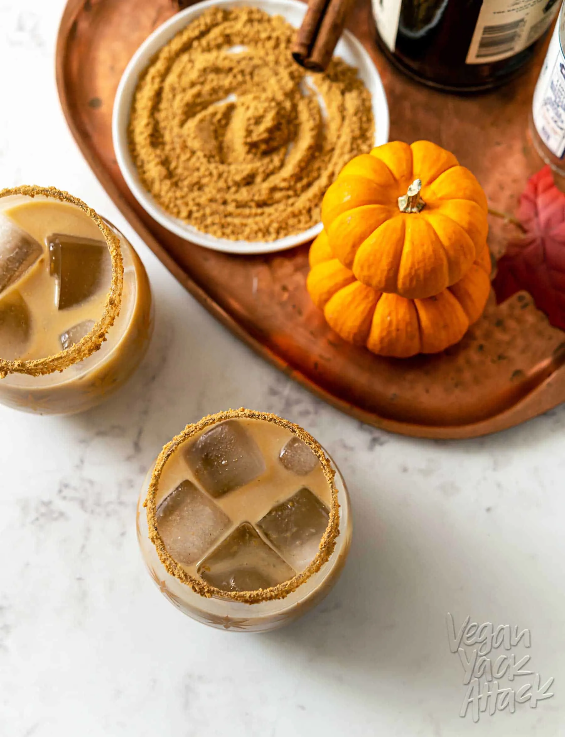 Two glasses filled with a tan-colored cocktail and pumpkins and crushed graham crackers around them
