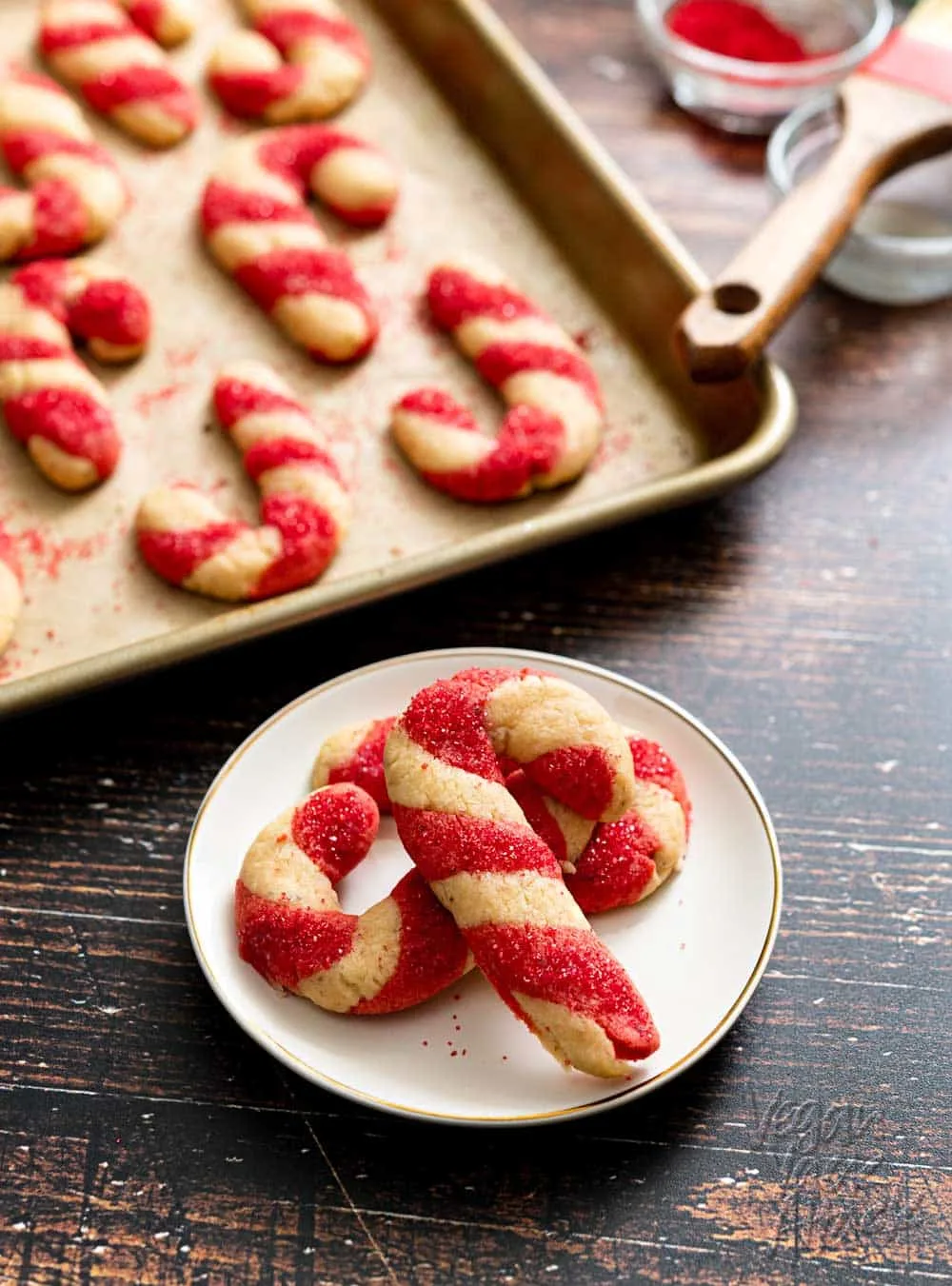 Candy cane-shaped cookies on a baking sheet next to colored sugar and pastry brush