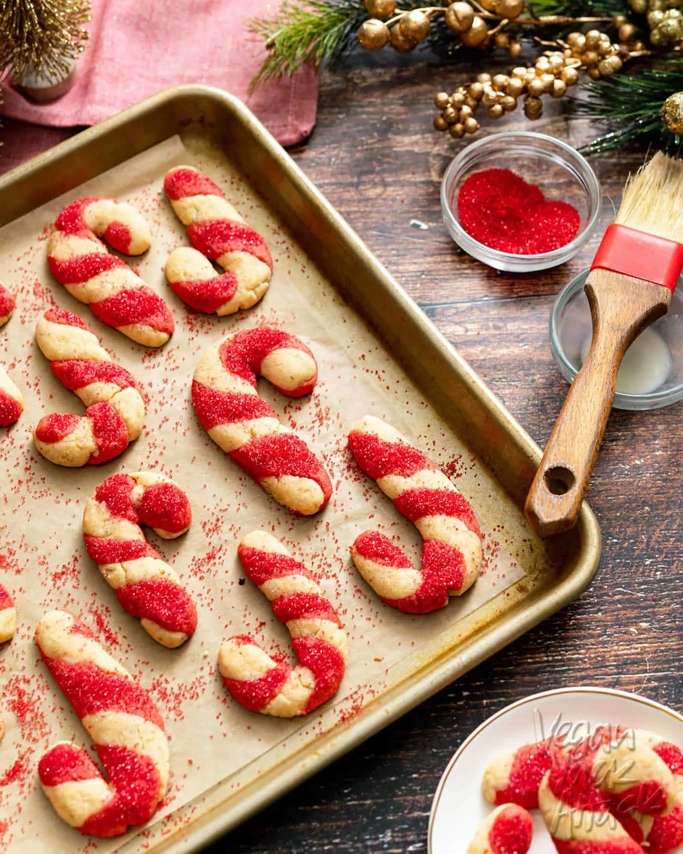 Candy cane-shaped cookies on a baking sheet next to colored sugar and pastry brush