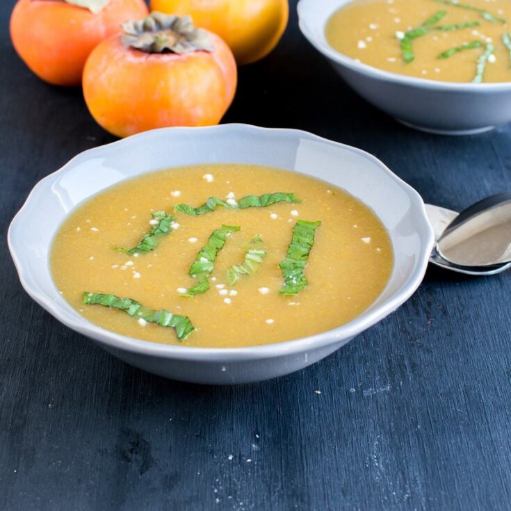 A front close view of persimmon habanero soup in a serving bowl with raw persimmon at the backdrop Print Recipe Persimmon Habanero Soup in a white bowl, garnished with basil