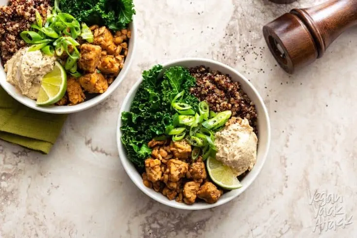 Two white bowls with kale, tempeh, quinoa, and bean dip on a marble background