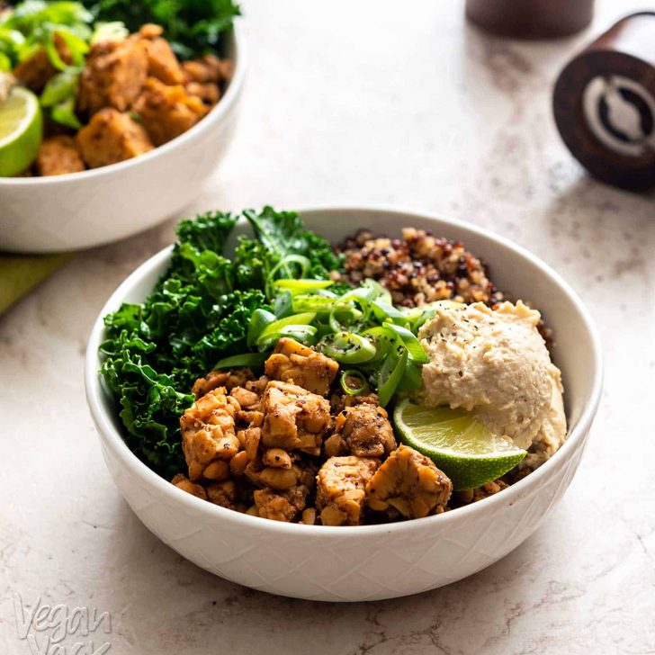 Two white bowls with kale, tempeh, quinoa, and bean dip on a marble background