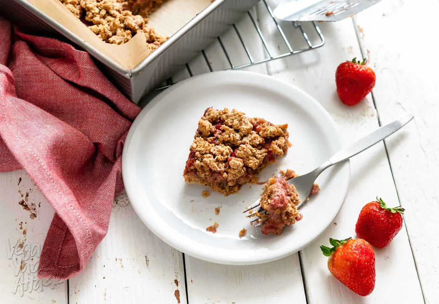 Piece of strawberry crumble bar cut out of a baking pan and bite on a fork next to it