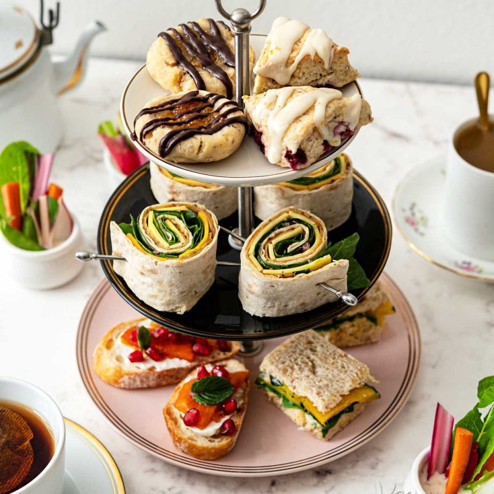 Plate tower filled with tea snacks including pinwheels, crostini, scones, cookies, and sandwiches