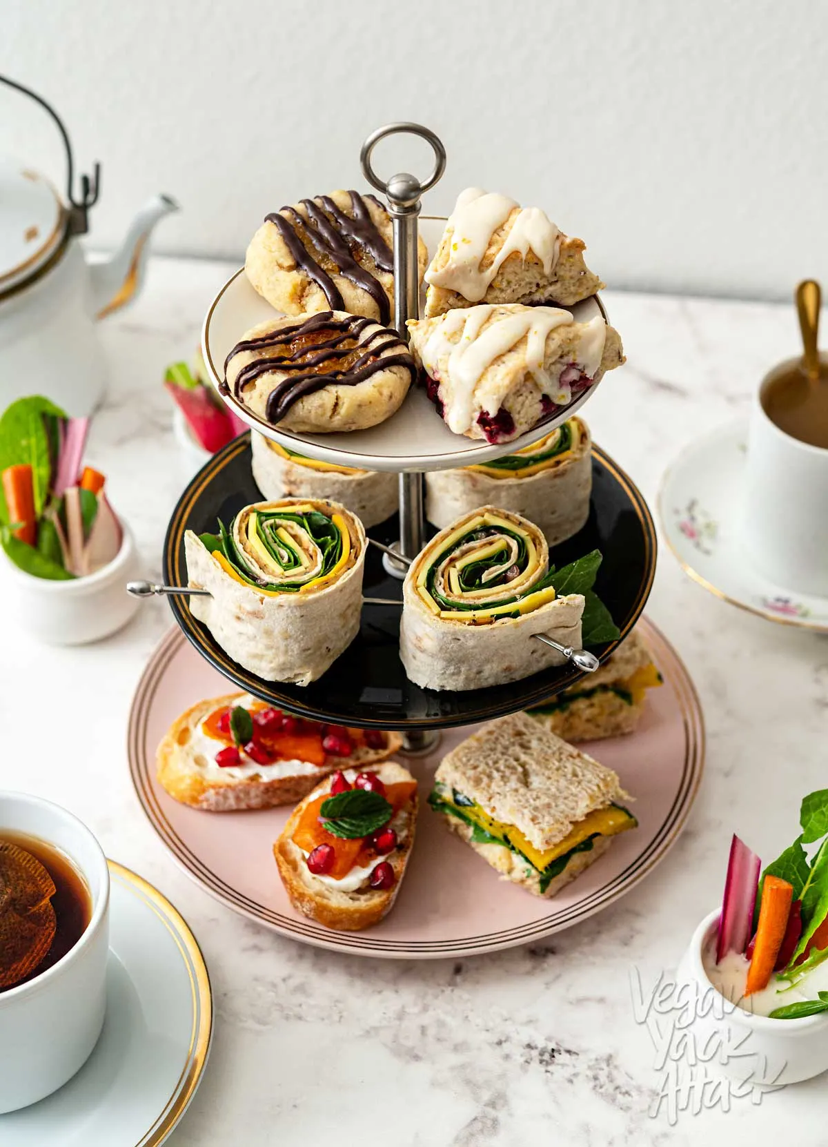 Plate tower filled with tea snacks including pinwheels, crostini, scones, cookies, and sandwiches