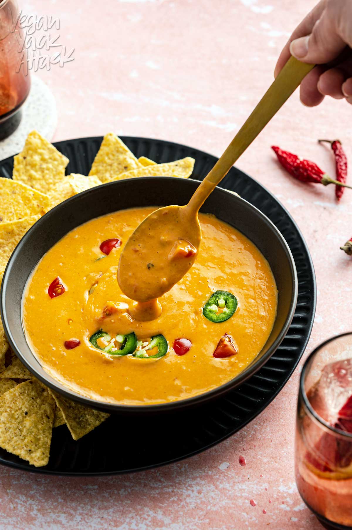 Bowl of bright orange queso dip on a plate with chips, on pink background with spoon scooping queso up