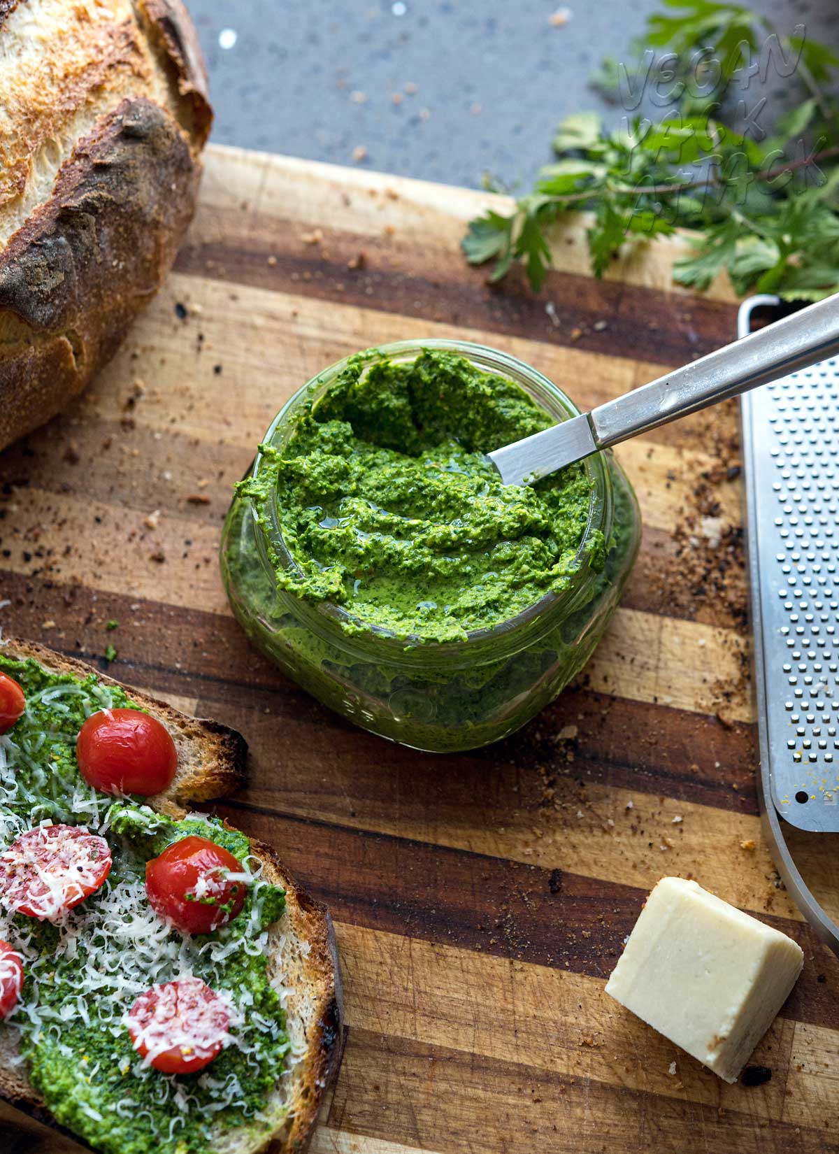 small jar of bright green pesto with knife in it on a cutting board next to tomato toast