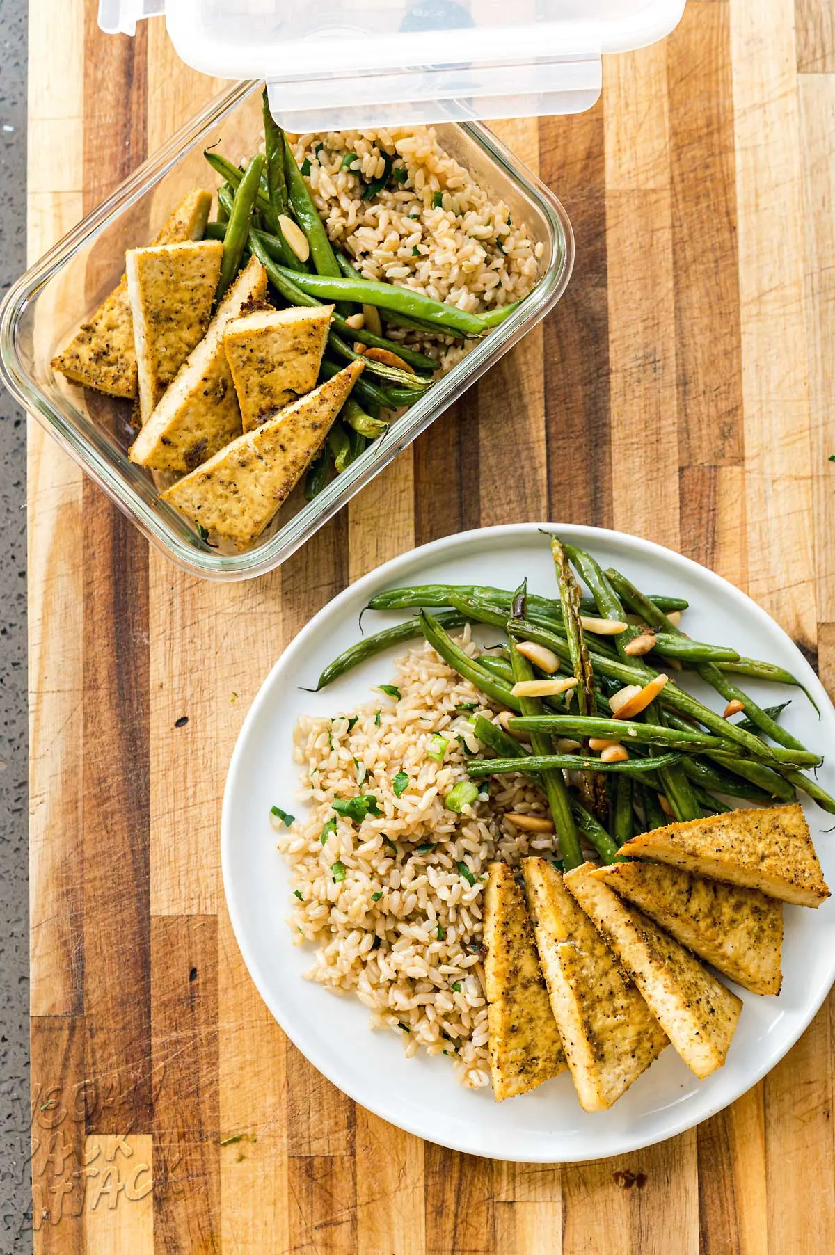 White plate of roasted tofu, green beans, and rice next to a container of the same on a cutting board