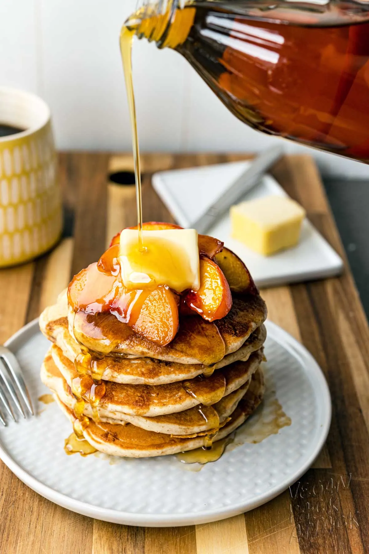 Stack of fluffy pancakes on a plate, topped with peaches, and being drizzled with maple syrup