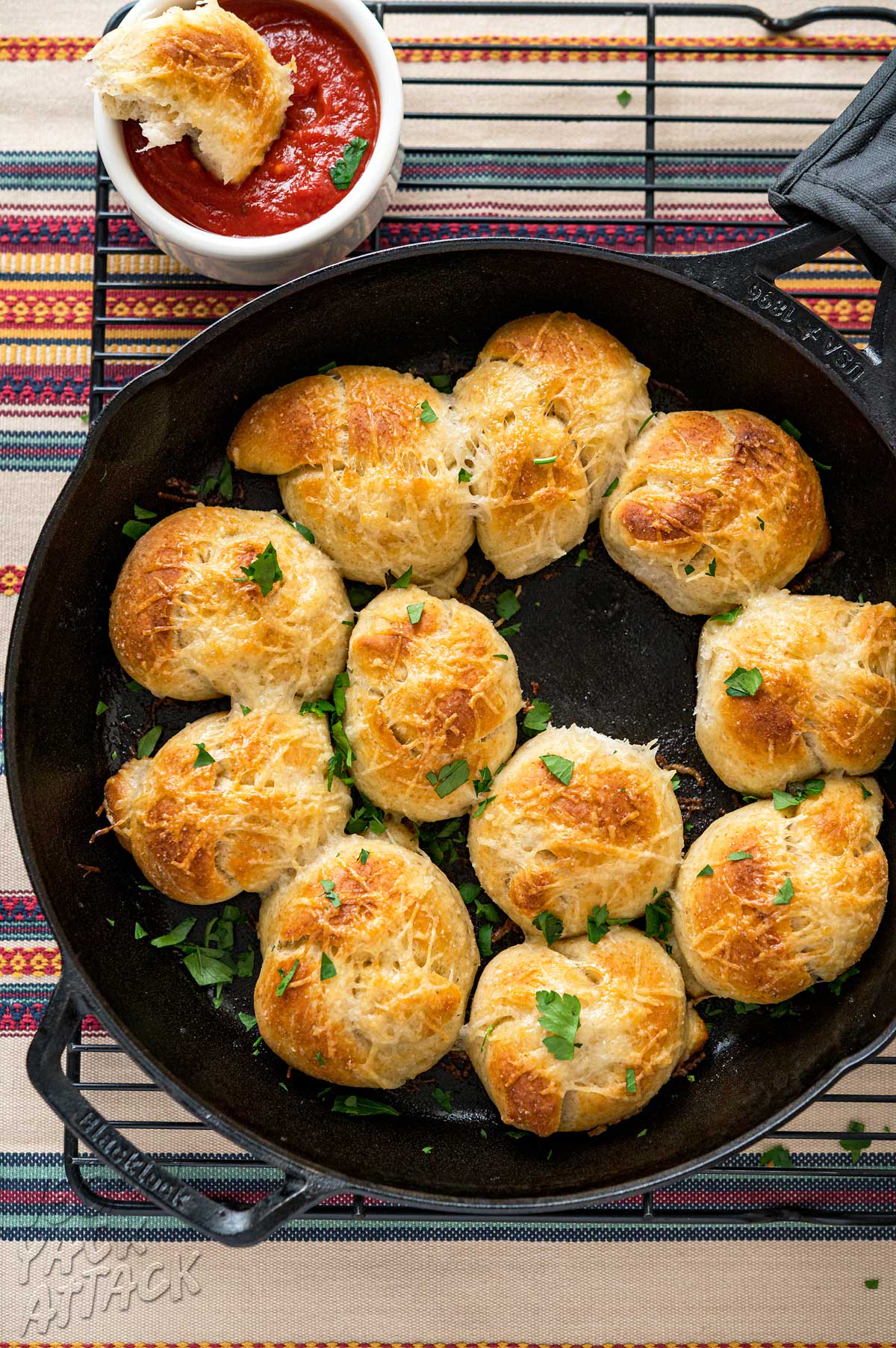 Garlic knots in a large cast iron pan on a cooling rack next to dipping sauce