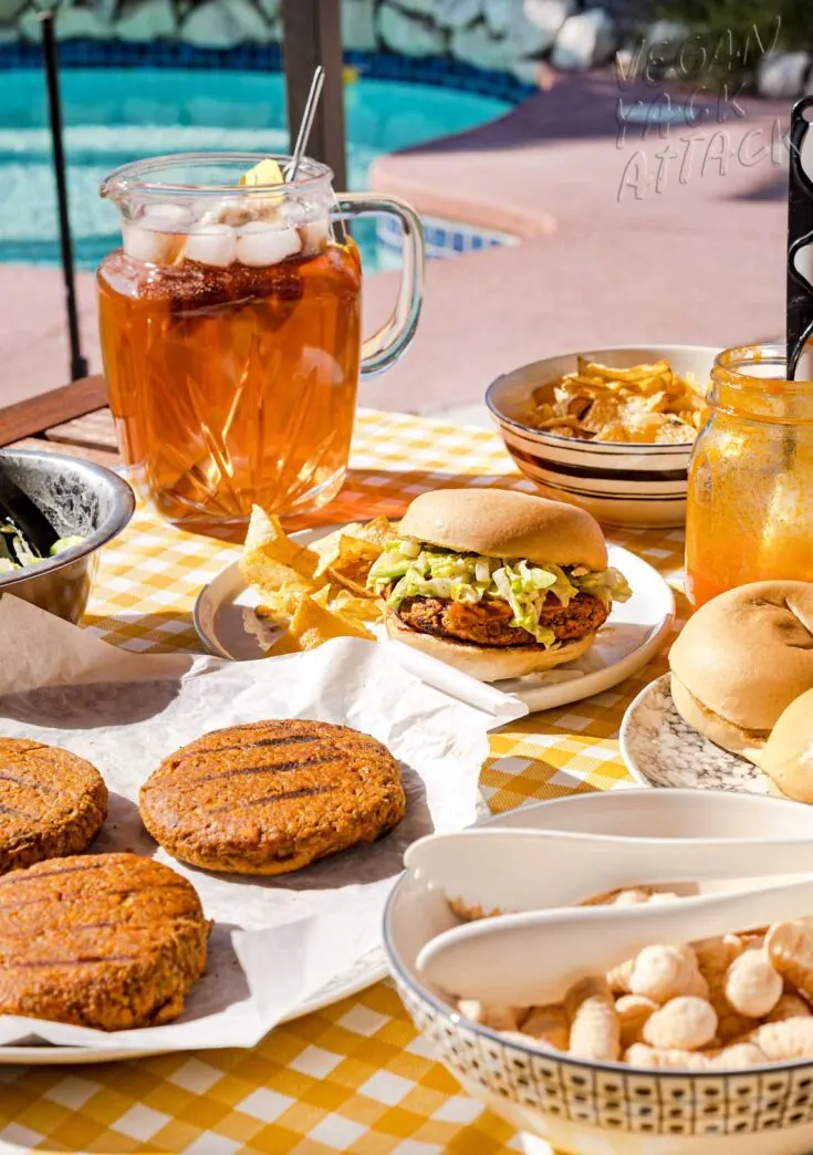 Buffalo Burger on a plate with chips, in a picnic table setting with pitcher of iced tea, plate of burger patties and bowl of chips