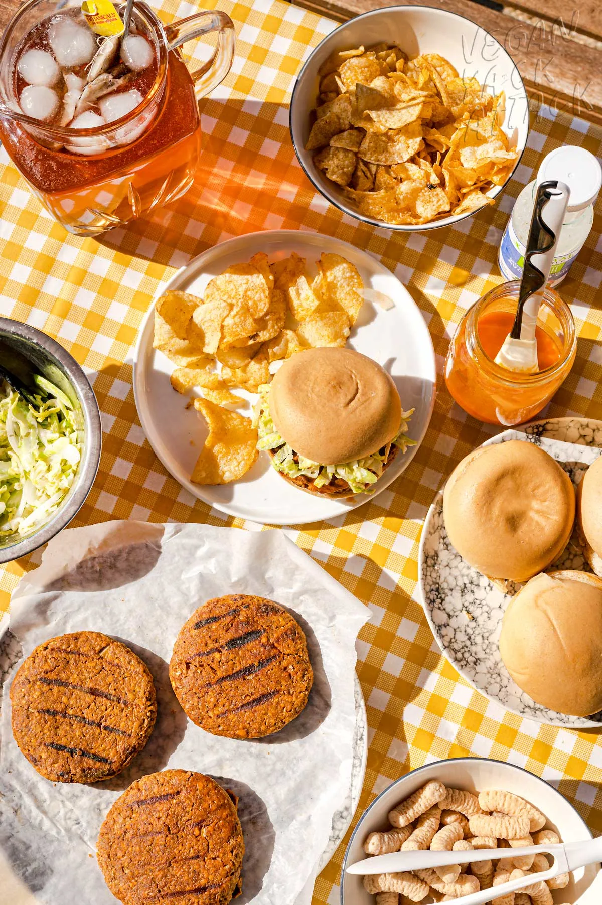 Buffalo Burger on a plate with chips, in a picnic table setting with pitcher of iced tea, plate of burger patties and bowl of chips
