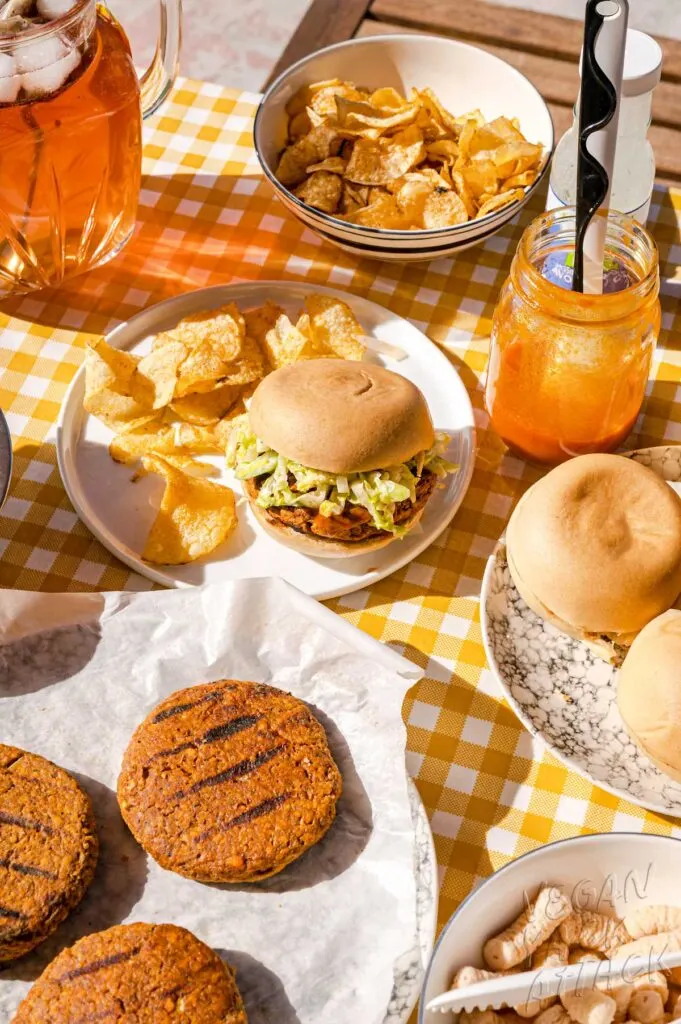 Buffalo Burger on a plate with chips, in a picnic table setting with pitcher of iced tea, plate of burger patties and bowl of chips