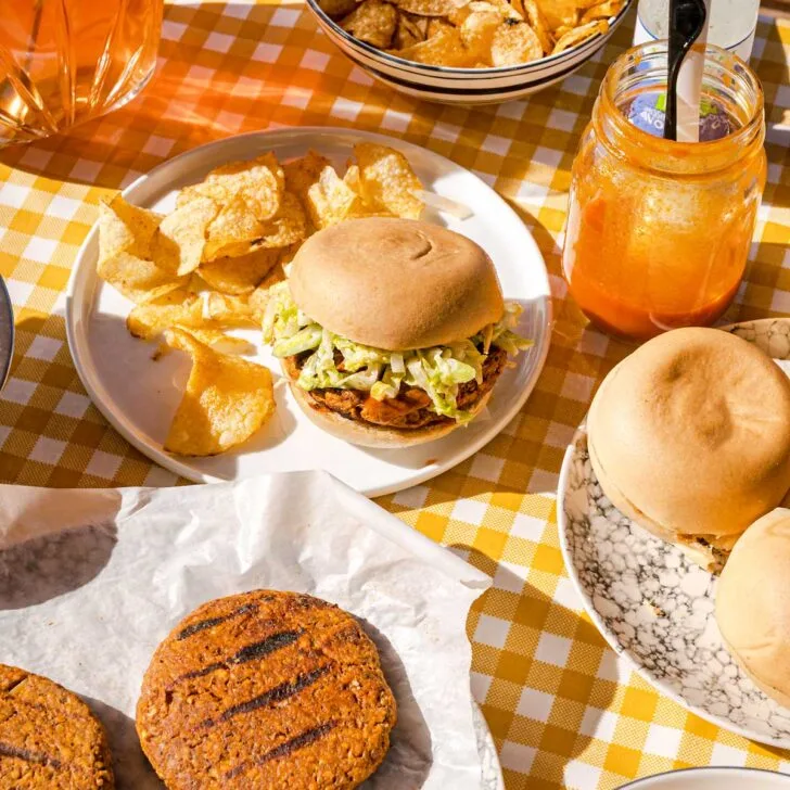 Buffalo Burger on a plate with chips, in a picnic table setting with pitcher of iced tea, plate of burger patties and bowl of chips