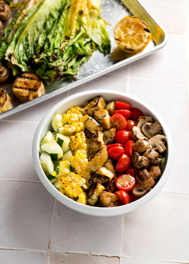 Large white bowl of salad with lines of chopped ingredients like cucumber, corn, eggplant, and tomato, next to a baking sheet with more ingredients